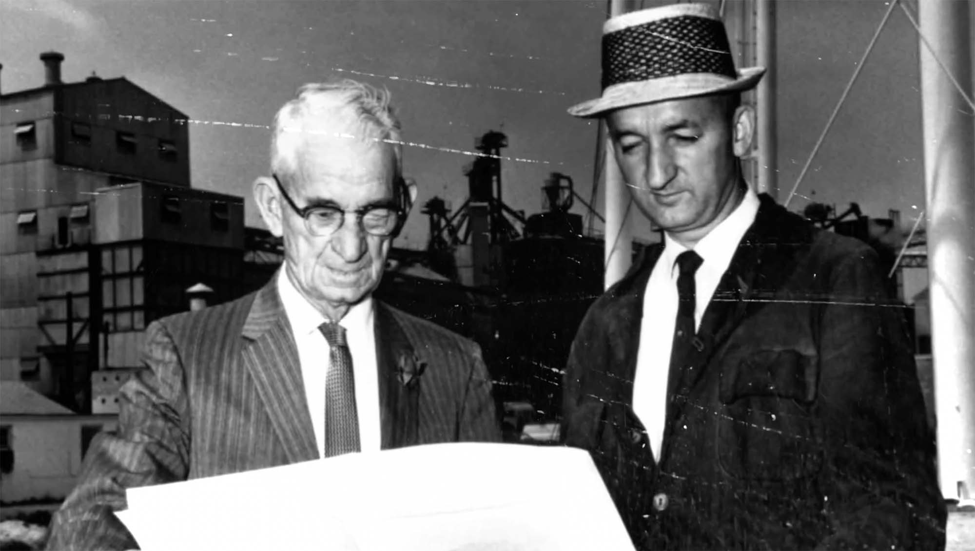 A vintage photo of two men reviewing a plan in front of a farm.